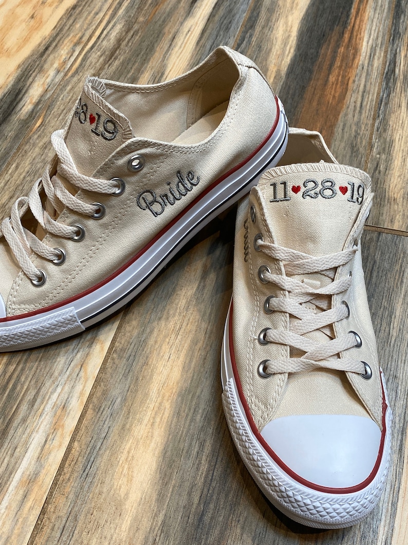 K&ouml;nnte beinhalten: Ein Paar cremefarbene Canvas-Sneaker mit rotem Besatz. Ein Schuh hat das Wort "Bride" bestickt, der andere das Datum "11.28.19".
