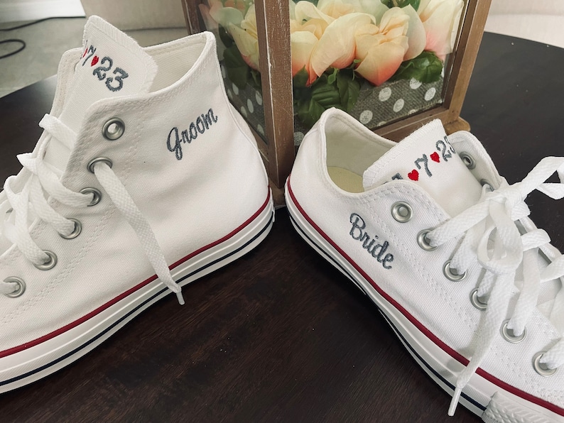 K&ouml;nnte beinhalten: Ein Paar wei&szlig;e Canvas-Sneaker mit roten Herzen und den Worten "Groom" und "Bride", die darauf gestickt sind. Die Sneaker haben einen roten und schwarzen Rand um die Sohle.