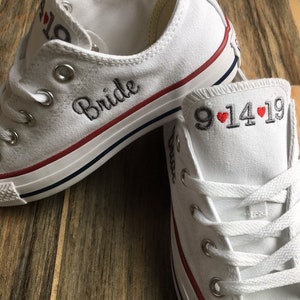 K&ouml;nnte beinhalten: Ein Paar wei&szlig;e Canvas-Schuhe mit rotem Besatz. Der linke Schuh hat das Wort "Bride" in Schwarz gestickt. Der rechte Schuh hat das Datum "9.14.19" in Schwarz mit zwei roten Herzen gestickt.