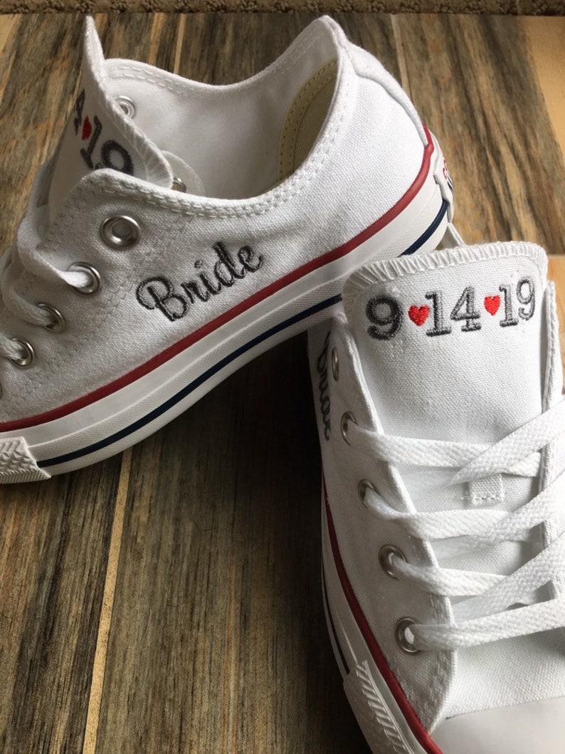 K&ouml;nnte beinhalten: Ein Paar wei&szlig;e Canvas-Schuhe mit rotem Besatz. Der linke Schuh hat das Wort "Bride" in Schwarz gestickt. Der rechte Schuh hat das Datum "9.14.19" in Schwarz mit zwei roten Herzen gestickt.