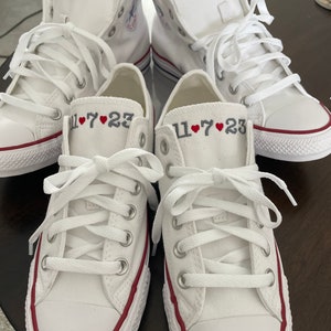K&ouml;nnte beinhalten: Vier wei&szlig;e Canvas-Sneaker mit rotem Besatz um die Sohle. Jeder Schuh hat das Datum "11&hearts;7&hearts;23" auf der Seite gestickt.