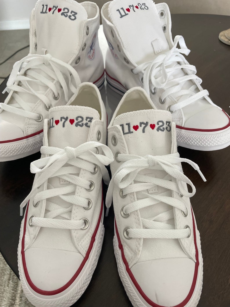 K&ouml;nnte beinhalten: Vier wei&szlig;e Canvas-Sneaker mit rotem Besatz um die Sohle. Jeder Schuh hat das Datum "11&hearts;7&hearts;23" auf der Seite gestickt.