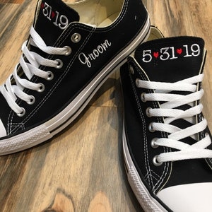 K&ouml;nnte beinhalten: Ein Paar schwarze Canvas-Sneaker mit wei&szlig;en Schn&uuml;rsenkeln. Der linke Schuh hat das Wort "Groom" an der Seite und das Datum "5.31.19" an der Spitze gestickt. Der rechte Schuh hat das Datum "5.31.19" mit roten Herzen an der Spitze.