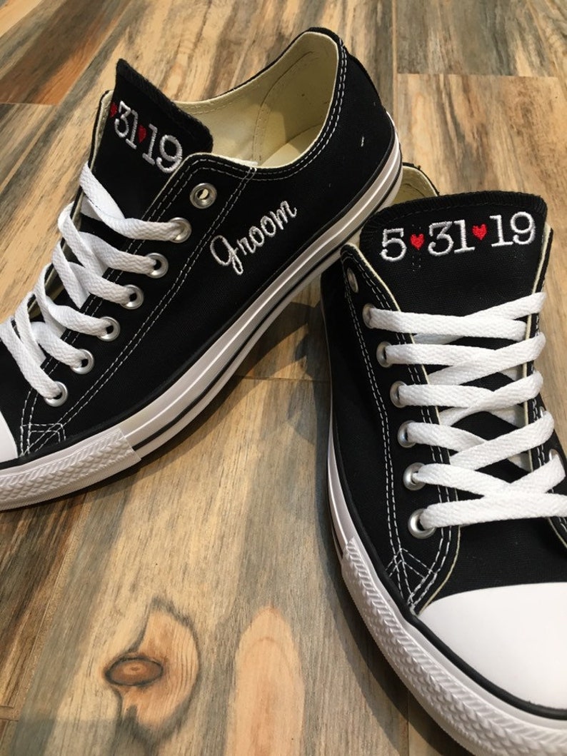 K&ouml;nnte beinhalten: Ein Paar schwarze Canvas-Sneaker mit wei&szlig;en Schn&uuml;rsenkeln. Der linke Schuh hat das Wort "Groom" an der Seite und das Datum "5.31.19" an der Spitze gestickt. Der rechte Schuh hat das Datum "5.31.19" mit roten Herzen an der Spitze.