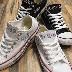 K&ouml;nnte beinhalten: Ein Paar wei&szlig;e Canvas-Sneaker mit rotem Besatz und dem Wort "Bride" auf einem Schuh gestickt. Der andere Schuh hat das Datum "5.31.19" gestickt. Ein Paar schwarze Canvas-Sneaker mit wei&szlig;en Schn&uuml;rsenkeln und dem Wort "Groom" auf einem Schuh gestickt. Der andere Schuh hat das Datum "5.31.19" gestickt.