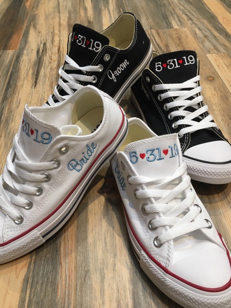 K&ouml;nnte beinhalten: Ein Paar wei&szlig;e Canvas-Sneaker mit rotem Besatz und dem Wort "Bride" auf einem Schuh gestickt. Der andere Schuh hat das Datum "5.31.19" gestickt. Ein Paar schwarze Canvas-Sneaker mit wei&szlig;en Schn&uuml;rsenkeln und dem Wort "Groom" auf einem Schuh gestickt. Der andere Schuh hat das Datum "5.31.19" gestickt.
