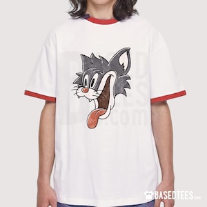 Puede incluir: Camiseta blanca con cuello y mangas ribeteados en rojo. La camiseta presenta un gráfico de gato de dibujos animados desgastado con la lengua fuera. El texto "BASEDTEES.com" está en la parte inferior de la camiseta.