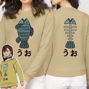 Puede incluir: Una sudadera de manga larga de color beige con un pez de dibujos animados en la parte delantera y un esqueleto de pez de dibujos animados en la parte posterior. El texto "うお" está impreso debajo de cada pez.