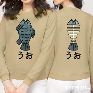 May include: A tan long-sleeved sweatshirt with a graphic of a blue fish on the front and a graphic of a fish skeleton on the back. The text "うお" is printed below each graphic.