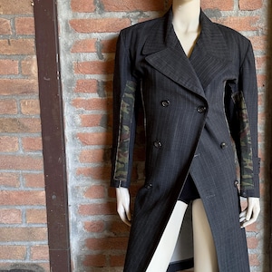 May include: A dark gray pinstripe double-breasted coat with camouflage patterned sleeves. The coat is displayed on a mannequin against a brick wall background. The coat has a long, asymmetrical design.