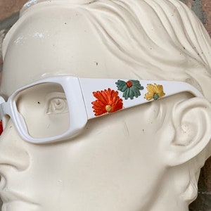 Könnte beinhalten: Weiße Kunststoff-Brille mit floralen Details in Orange, Grün und Gelb.
