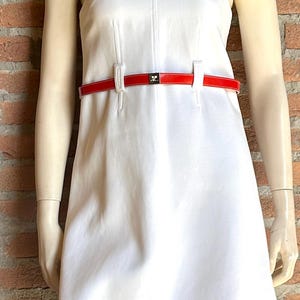 Puede incluir: Un vestido blanco sin mangas con cintur&oacute;n rojo. El vestido tiene una silueta sencilla en forma de A y un cuello alto. El cintur&oacute;n rojo ci&ntilde;e la cintura. El vestido se exhibe en un maniqu&iacute;.