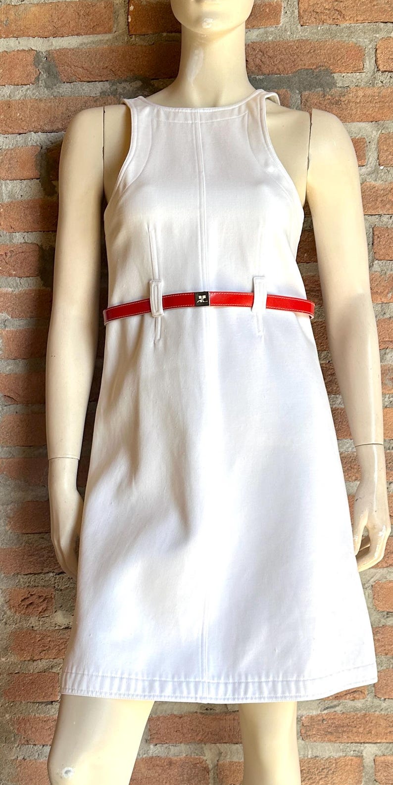Puede incluir: Un vestido blanco sin mangas con cintur&oacute;n rojo. El vestido tiene una silueta sencilla en forma de A y un cuello alto. El cintur&oacute;n rojo ci&ntilde;e la cintura. El vestido se exhibe en un maniqu&iacute;.