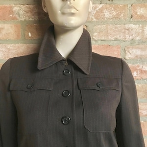 Può includere: Giacca marrone a gessato con ampio colletto, due tasche sul petto e bottoni neri. La giacca è esposta su un manichino contro un muro di mattoni.