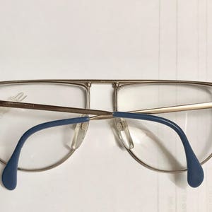 May include: Gold-rimmed eyeglasses with a double bridge and blue temples. The lenses are clear, and the glasses have a classic aviator style. The frame is a light gold color.
