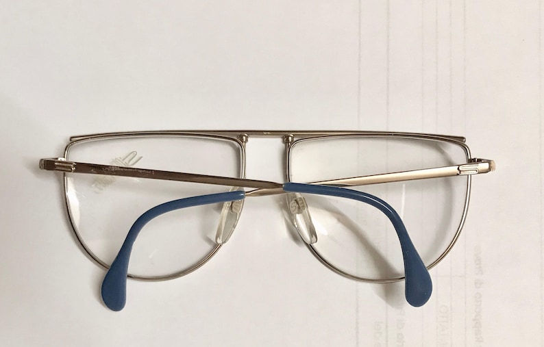 May include: Gold-rimmed eyeglasses with a double bridge and blue temples. The lenses are clear, and the glasses have a classic aviator style. The frame is a light gold color.