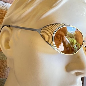 May include: Sunglasses featuring oval-shaped amber-tinted lenses and a metallic frame. The side panels of the frame display a distinctive ribbed, gold-toned texture that extends from the lens rim toward the thin, dark-colored temple arms.