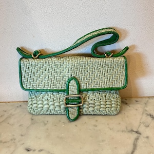 Può includere: Una piccola borsa a mano intrecciata nei toni del verde. La borsa ha una forma rettangolare con chiusura a patta e manico. I bordi e il manico sono rifiniti in un verde più scuro e la chiusura è color oro.