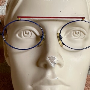 May include: A white mannequin head wearing round blue-rimmed glasses with a red top bar. The glasses have gold accents on the sides.