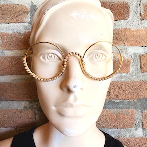 May include: Gold-toned circular spectacles featuring a thin metal frame adorned with a continuous row of small, clear, sparkling rhinestones along the outer edges of the lenses. The glasses have a high bridge and simple, slender metal temples.