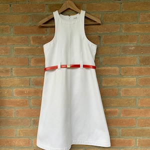 Puede incluir: Vestido blanco sin mangas con cintur&oacute;n rojo. El vestido est&aacute; colgado de una percha de madera contra una pared de ladrillos. El cintur&oacute;n rojo tiene una hebilla dorada.