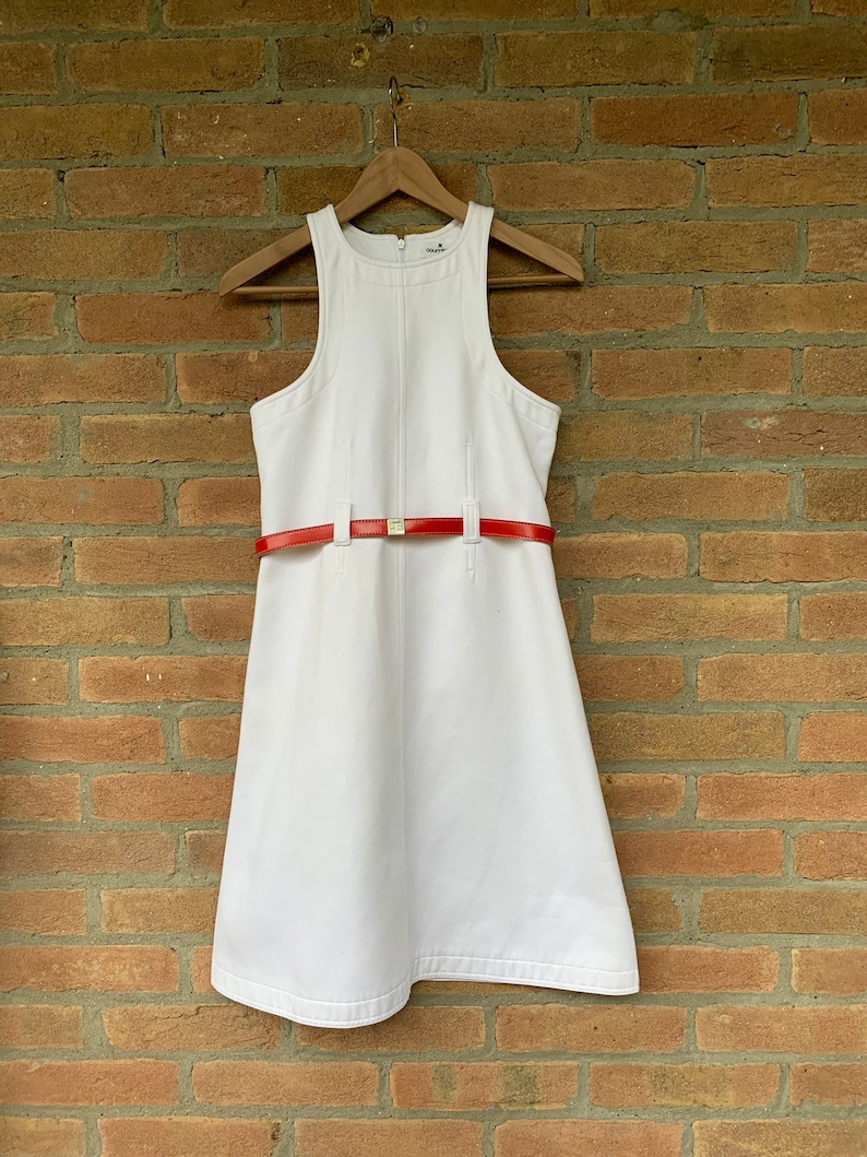 Puede incluir: Vestido blanco sin mangas con cintur&oacute;n rojo. El vestido est&aacute; colgado de una percha de madera contra una pared de ladrillos. El cintur&oacute;n rojo tiene una hebilla dorada.