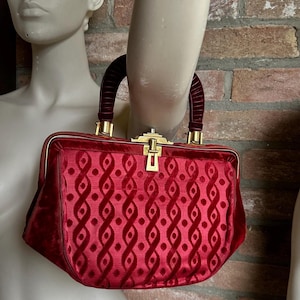 Pu&ograve; includere: Una borsa a mano in velluto rosso intenso con un motivo geometrico ripetitivo di linee ondulate verticali e punti. La borsa presenta una struttura rigida, un manico arcuato in velluto e una chiusura a scatto in metallo dorato sulla parte superiore.