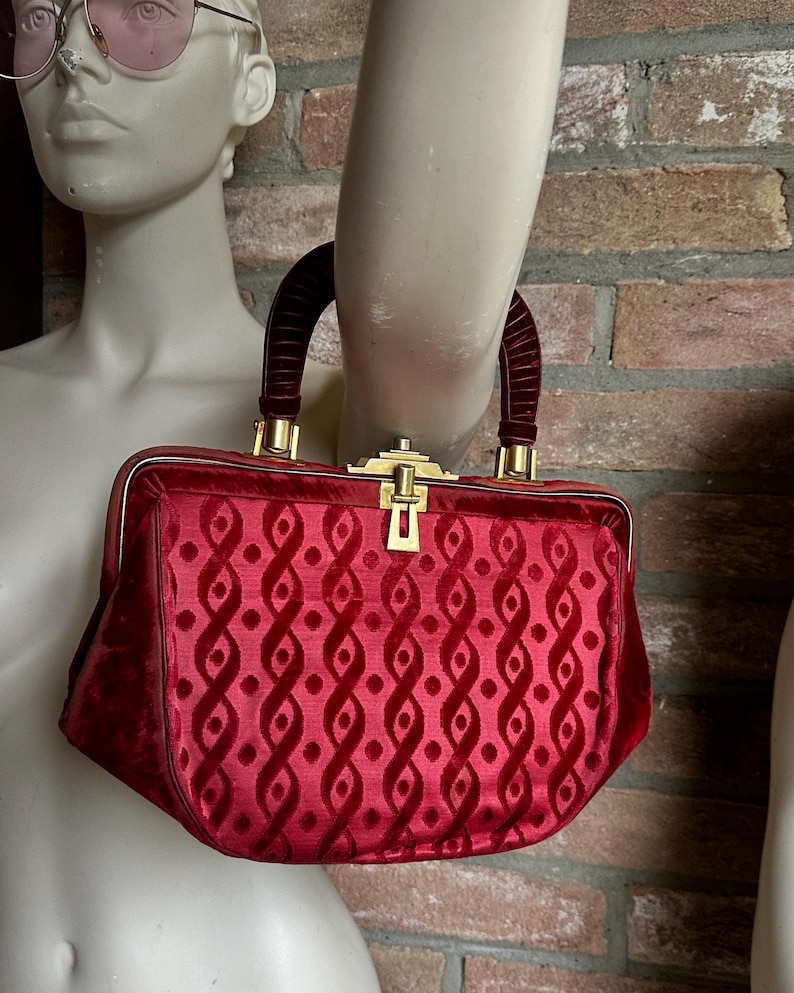 Pu&ograve; includere: Una borsa a mano in velluto rosso intenso con un motivo geometrico ripetitivo di linee ondulate verticali e punti. La borsa presenta una struttura rigida, un manico arcuato in velluto e una chiusura a scatto in metallo dorato sulla parte superiore.