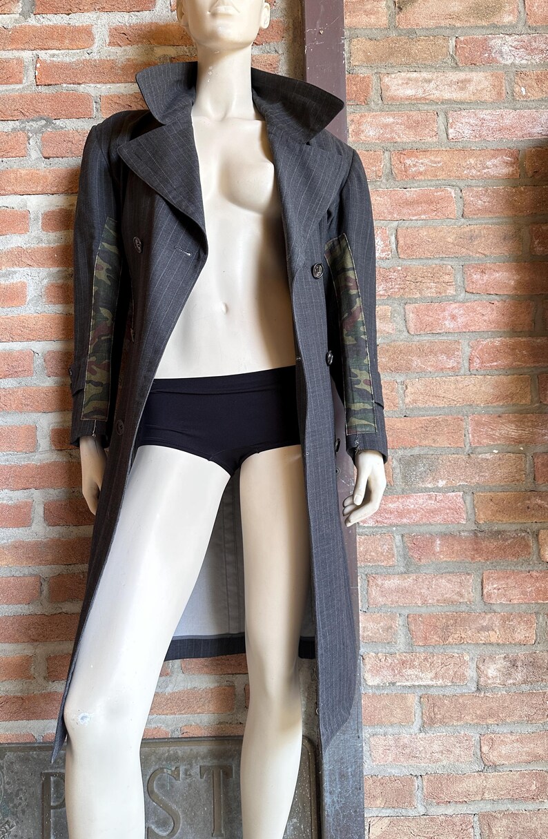 May include: A gray pinstripe trench coat with camouflage patterned sleeves is displayed on a mannequin. The coat is buttoned and has a wide collar. The mannequin is also wearing black underwear.