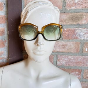 May include: Oversized sunglasses featuring a thick, amber-colored plastic frame with a distinct angular top edge. The lenses exhibit a gradient tint, transitioning from a lighter green hue at the top to a darker, smoky shade toward the bottom rim.