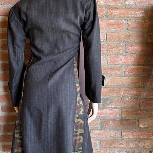 May include: A dark gray pinstripe coat with long sleeves and a contrasting camouflage pattern down the sides. The coat is displayed on a mannequin against a brick wall.