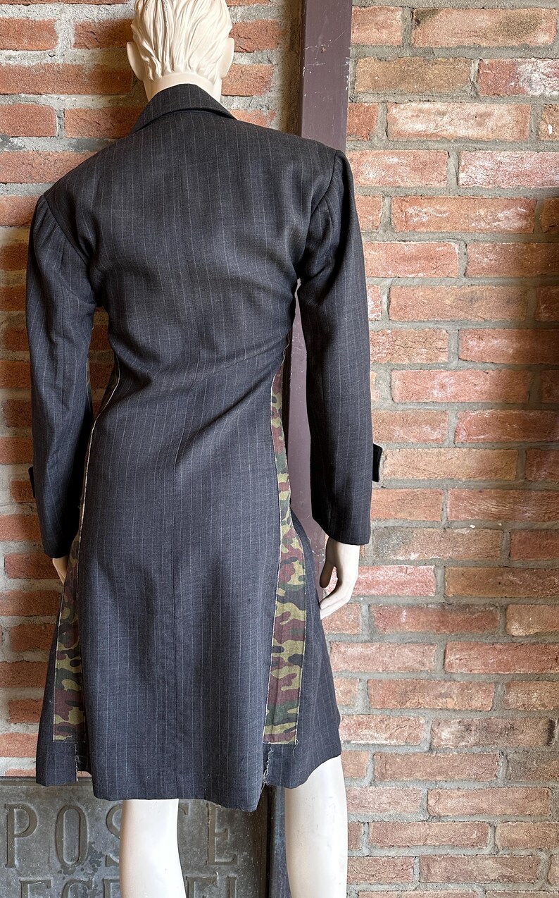 May include: A dark gray pinstripe coat with long sleeves and a contrasting camouflage pattern down the sides. The coat is displayed on a mannequin against a brick wall.