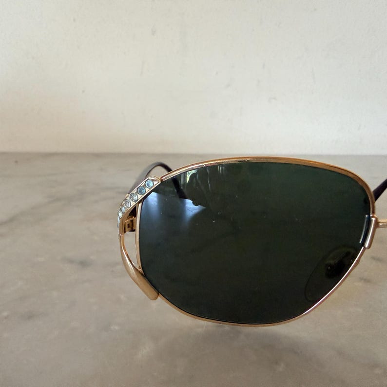 May include: Sunglasses with a gold-toned metal frame and dark green lenses. The outer edge of the frame features a decorative accent set with small, clear, sparkling crystals. The design includes a thin metal bridge and dark temple arms.
