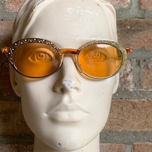 Op de afbeelding: Ovale zonnebril met doorschijnende oranje glazen en goudkleurige metalen monturen. De randen zijn versierd met een herhalend geometrisch patroon en kleine ingebedde heldere kristallen. De brug en de pootjes zijn gemaakt van bijpassend oranje materiaal.