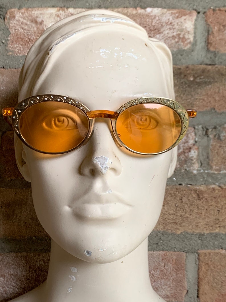 Op de afbeelding: Ovale zonnebril met doorschijnende oranje glazen en goudkleurige metalen monturen. De randen zijn versierd met een herhalend geometrisch patroon en kleine ingebedde heldere kristallen. De brug en de pootjes zijn gemaakt van bijpassend oranje materiaal.