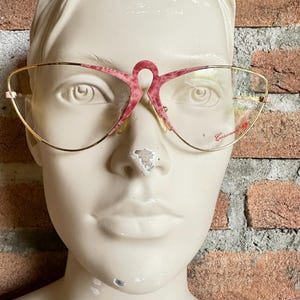 Puede incluir: Un par de gafas de ojo de gato de color rosa y dorado con un diseño de puente único. Las gafas están etiquetadas como "Casanova".