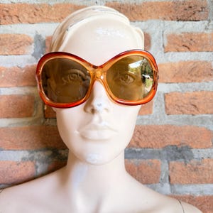 Può includere: Occhiali da sole oversize con una montatura in plastica arancione ambra traslucida dalla forma arrotondata ispirata alle farfalle. Le lenti sono colorate in una tonalità marrone calda, offrendo una visione chiara attraverso le lenti.