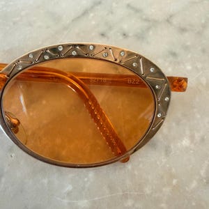 Op de afbeelding: Ovale zonnebril met oranje getinte glazen en een goudkleurig metalen montuur. Het montuur is versierd met kleine, heldere ingebedde kristallen en ge&euml;tste geometrische lijnen. De binnenkant van het glas is gemarkeerd met de tekst 52718 BZ2.