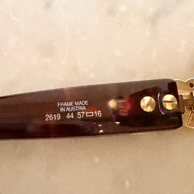 May include: A dark brown tortoiseshell eyeglass temple arm featuring gold-tone metal screws. The interior text reads FRAME MADE IN AUSTRIA 2619 44 57 16. The material has a glossy finish and the hinge mechanism is visible on the right side of the frame.