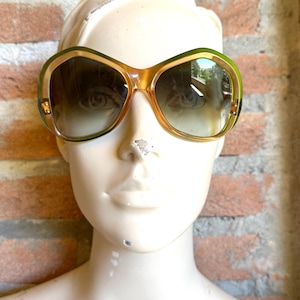 Può includere: Occhiali da sole vintage oversize con montatura marrone chiaro e lenti sfumate. Le montature hanno un accento verde sul bordo superiore. Gli occhiali da sole sono esposti su una testa di manichino contro un muro di mattoni.