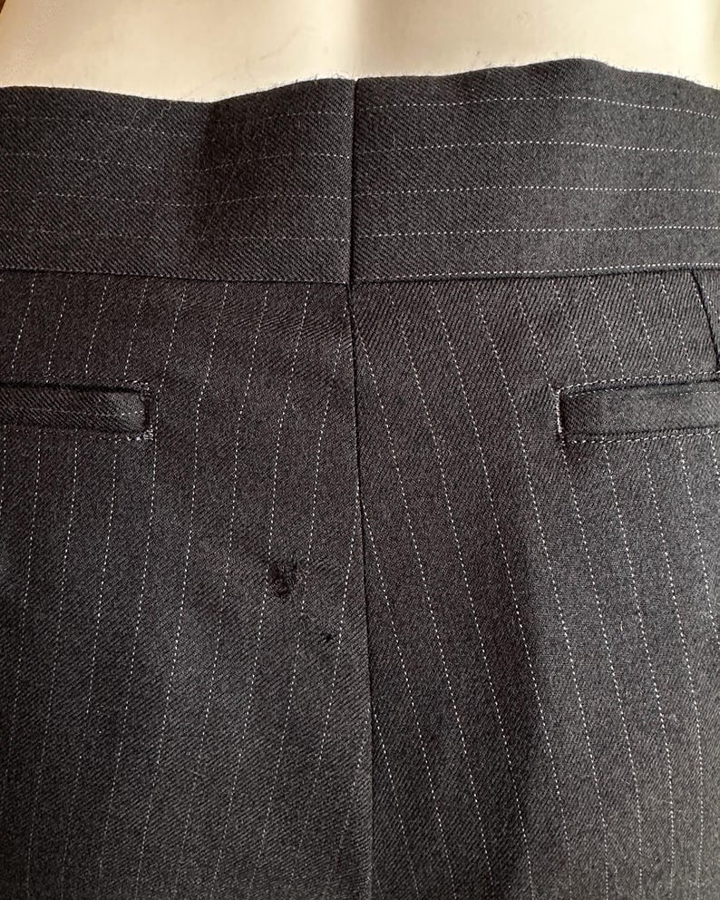 Peut inclure: Gros plan du dos d'un pantalon &agrave; fines rayures gris fonc&eacute;. Le tissu pr&eacute;sente des rayures blanches verticales. Le pantalon a deux poches arri&egrave;re et une couture au centre. La ceinture est visible en haut.