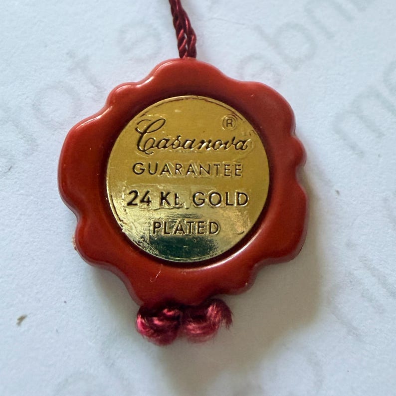 Op de afbeelding: Een cirkelvormig goudkleurig metalen label met een rode wasachtige zegelrand. De tekst op het label luidt Casanova, GUARANTEE, 24 Kt. GOLD, PLATED. Een dun rood gevlochten koord is aan de boven- en onderkant van het zegel bevestigd.