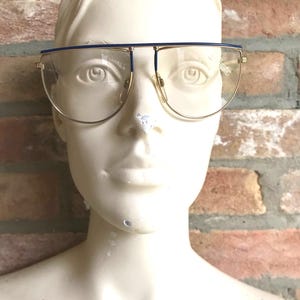 May include: Gold-rimmed eyeglasses with a blue top bar and clear lenses. The glasses have a rectangular shape and are set against a brick wall background. The word "quality" is visible on the lens.
