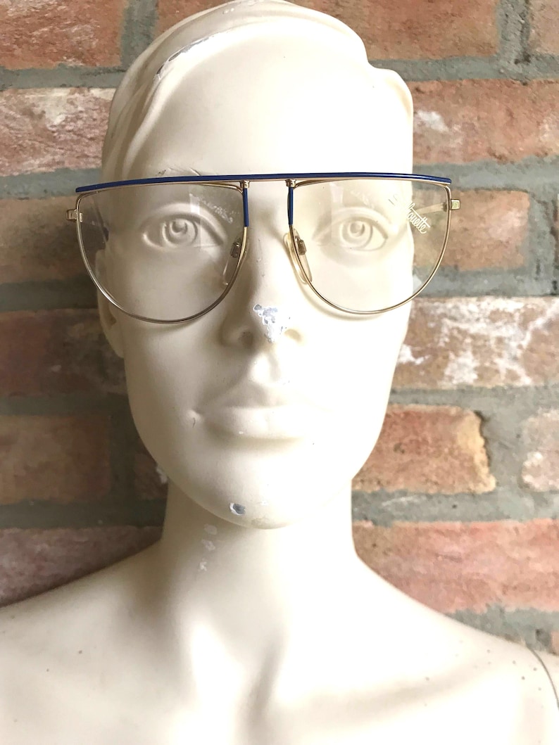 May include: Gold-rimmed eyeglasses with a blue top bar and clear lenses. The glasses have a rectangular shape and are set against a brick wall background. The word "quality" is visible on the lens.