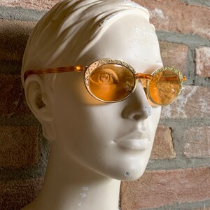 Op de afbeelding: Ovale zonnebril met amberkleurige glazen en goudkleurige metalen monturen met een ge&euml;tst geometrisch patroon langs de randen. De pootjes zijn gemaakt van een doorschijnend bruin materiaal met een schildpadpatroon.