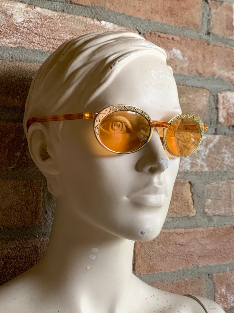 Op de afbeelding: Ovale zonnebril met amberkleurige glazen en goudkleurige metalen monturen met een ge&euml;tst geometrisch patroon langs de randen. De pootjes zijn gemaakt van een doorschijnend bruin materiaal met een schildpadpatroon.