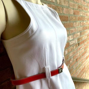 Puede incluir: Un vestido blanco sin mangas con cintur&oacute;n rojo. El vestido tiene un dise&ntilde;o &uacute;nico con cortes en los hombros y una silueta ajustada. El cintur&oacute;n rojo resalta la cintura. El cintur&oacute;n tiene una hebilla plateada. El vestido est&aacute; hecho de una tela suave. El vestido mide unos 100 cm de largo.