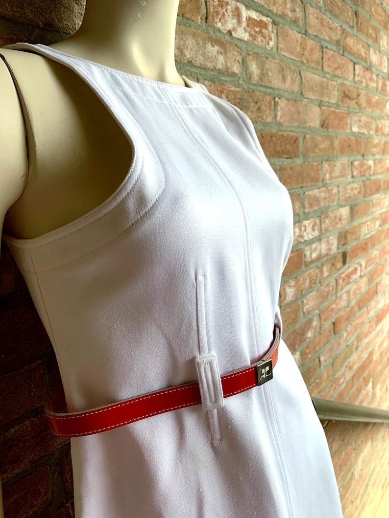 Puede incluir: Un vestido blanco sin mangas con cintur&oacute;n rojo. El vestido tiene un dise&ntilde;o &uacute;nico con cortes en los hombros y una silueta ajustada. El cintur&oacute;n rojo resalta la cintura. El cintur&oacute;n tiene una hebilla plateada. El vestido est&aacute; hecho de una tela suave. El vestido mide unos 100 cm de largo.