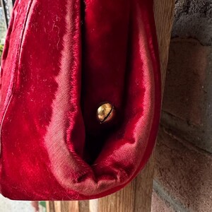 Pu&ograve; includere: Un oggetto in tessuto di velluto rosso brillante con una consistenza morbida e felpata, dotato di un piccolo campanello rotondo in metallo dorato fissato sul lato. Il tessuto presenta cuciture visibili e una tonalit&agrave; cremisi profonda.