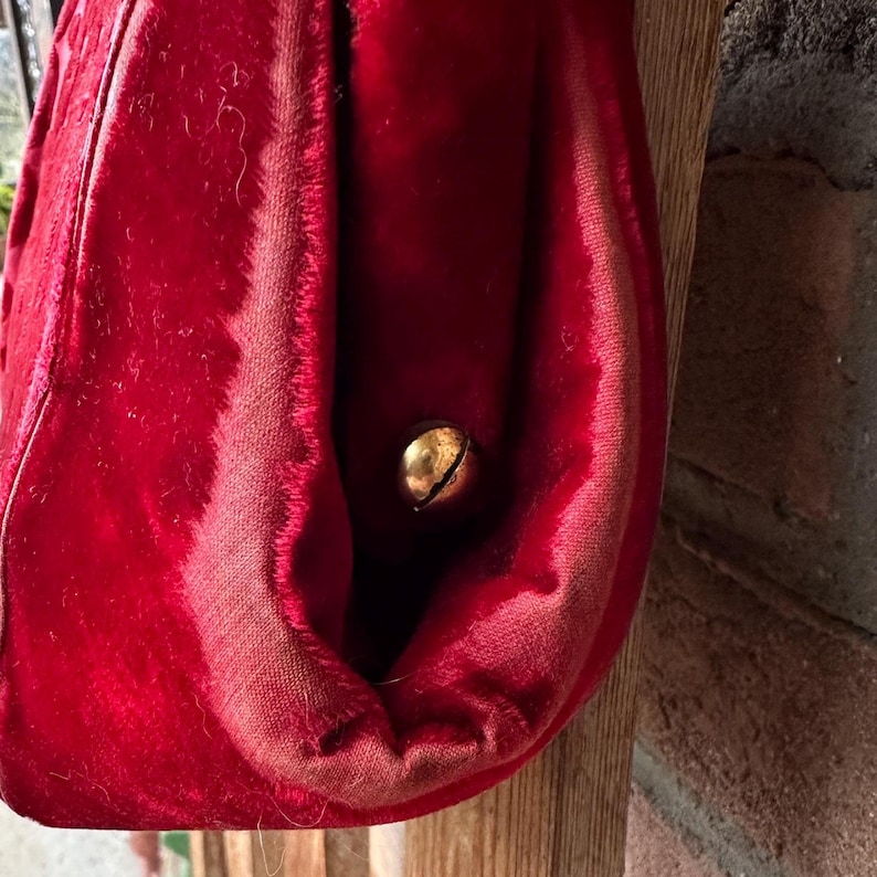 Pu&ograve; includere: Un oggetto in tessuto di velluto rosso brillante con una consistenza morbida e felpata, dotato di un piccolo campanello rotondo in metallo dorato fissato sul lato. Il tessuto presenta cuciture visibili e una tonalit&agrave; cremisi profonda.